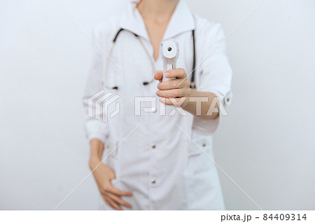Temperature measurement gun in doctor hands. Close-up shot of doctor wearing protective surgical mask ready to use infrared isometric thermometer gun to check body temperature for virus symptoms. Temperature measurement gun in doctor hands. Close-up shot of doctor wearing protective surgical mask ready to use infrared isometric thermometer gun to check body temperature for virus symptoms. 84409314