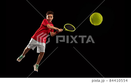Full-length portrait of boy, tennis player in motion, kicking ball with racket isolated over black backgound Full-length portrait of boy, tennis player in motion, kicking ball with racket isolated over black backgound 84410554