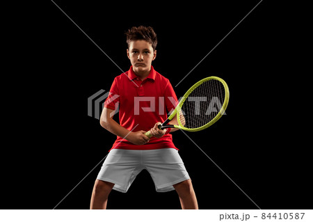 Cropped portrait of boy, tennis player in uniform with racket posing isolated over black background 84410587
