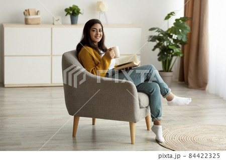 Weekend pastime. Happy arab woman reading book, drinking coffee and smiling at camera, sitting in comfy armchair Weekend pastime. Happy arab woman reading book, drinking coffee and smiling at camera, sitting in comfy armchair 84422325