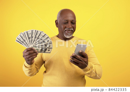Cheerful senior black man with money and cellphone winning lottery or casino bet on orange studio background Cheerful senior black man with money and cellphone winning lottery or casino bet on orange studio background 84422335