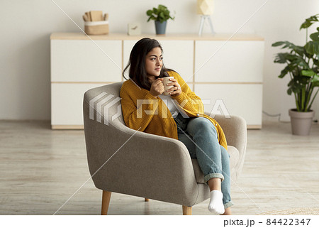 Positive arab lady sitting in armchair and enjoying morning coffee, warming hands on cup of hot beverage Positive arab lady sitting in armchair and enjoying morning coffee, warming hands on cup of hot beverage 84422347