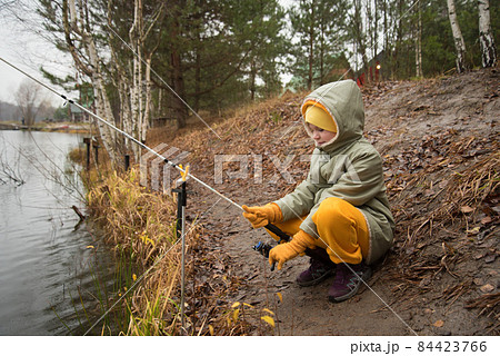 The child is a fisherman by the autumn lake. A child in warm clothes in the rain sits on the shore with a fishing rod The child is a fisherman by the autumn lake. A child in warm clothes in the rain sits on the shore with a fishing rod 84423766