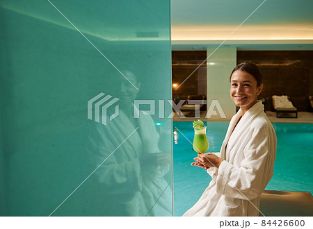 Middle aged pretty woman in white bathrobe stands by the pool in a wellness spa centre, holds freshly squeezed vitamin juice in hands, smiles looking at camera. Healthy lifestyle, recreation concept Middle aged pretty woman in white bathrobe stands by the pool in a wellness spa centre, holds freshly squeezed vitamin juice in hands, smiles looking at camera. Healthy lifestyle, recreation concept 84426600