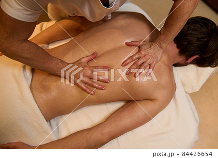 Overhead view of unrecognizable professional masseuse doing therapeutic healing massage of back muscles of a young man, relaxing on massage table at wellness spa centre. Alternative therapy concept Overhead view of unrecognizable professional masseuse doing therapeutic healing massage of back muscles of a young man, relaxing on massage table at wellness spa centre. Alternative therapy concept 84426654