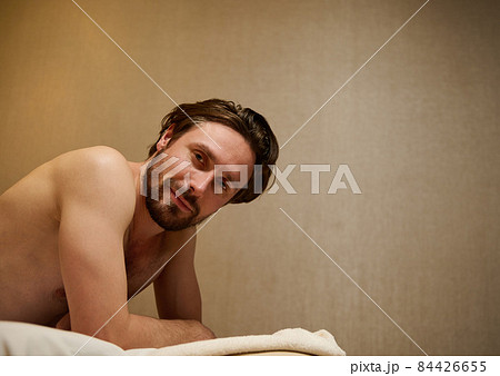 Handsome middle aged Caucasian man lying on a massage table, smiles looking at camera, getting ready to receive professional body massage therapy at wellness center. Ayurveda and body care concept 84426655