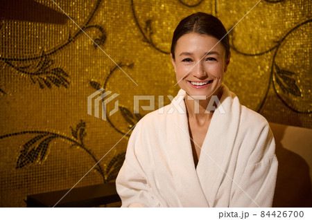 Close-up portrait of a beautiful stunning delighted middle aged European woman in white terry bathrobe smiling toothy smile looking at camera, sitting on an armchair in wellness spa centre lounge area Close-up portrait of a beautiful stunning delighted middle aged European woman in white terry bathrobe smiling toothy smile looking at camera, sitting on an armchair in wellness spa centre lounge area 84426700