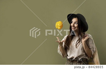 Smiling brunette young woman in a beige t-shirt and a hat holding a reusable mesh bag with fresh vegetables and fruits on a shoulder and throwing a yellow pepper in a air. Zero waste concept. 84428935