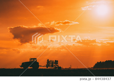 Tractor Rides On Countryside Road. Beginning Of Agricultural Spring Season. Cultivator Pulled By A Tractor In Rural Field Landscape Under Sunny Summer Sunset Sunrise Sky. Backlit Dramatic Lighting 84438437