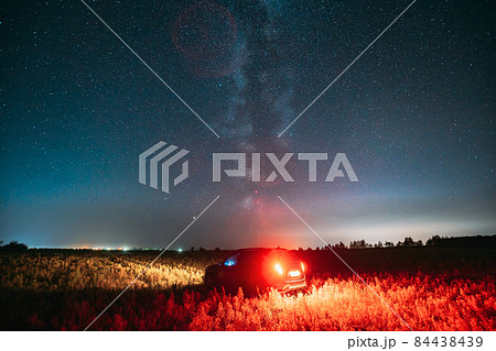 Night Starry Sky With Glowing Stars Above Countryside Road Landscape With SUV Car Vehicle Automobile. Milky Way Galaxy And Rural Field Meadow 84438439