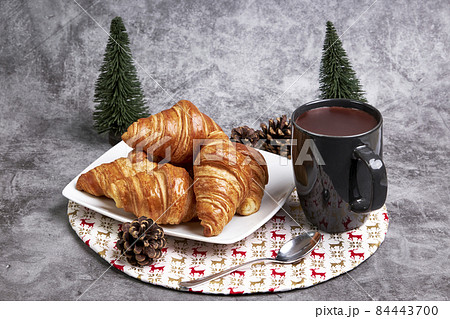 Cup of hot chocolate with a plate with croissant on a Christmas tablecloth and some Christmas fir trees and pine cones Cup of hot chocolate with a plate with croissant on a Christmas tablecloth and some Christmas fir trees and pine cones 84443700