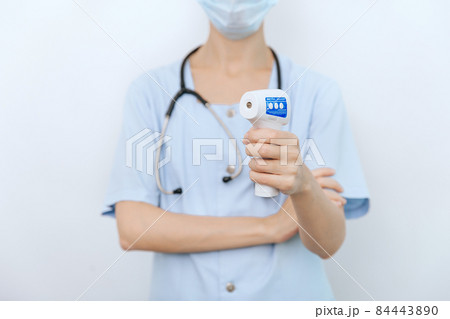 Temperature measurement gun in doctor hands. Close-up shot of doctor wearing protective surgical mask ready to use infrared isometric thermometer gun to check body temperature for virus symptoms. 84443890