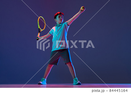 Full-length portrait of boy, tennis player in motion, kicking ball with racket isolated over purple background in neon 84445164