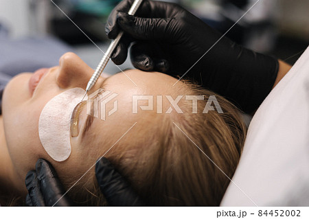 Close-up of eyelash care treatment procedures. Beautiful female, blond hair model have eyelashes lamination. Woman doing staining, curling, laminating and extension for lashes 84452002