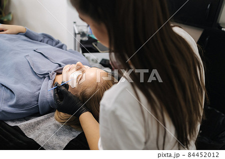 Woman doing eyelashes lamination in beauty studio. Blond hair model has staining, curling, laminating and extension for lashes. Black gloves 84452012