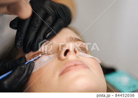 Close-up of eyelash care treatment procedures. Beautiful female, blond hair model have eyelashes lamination. Woman doing staining, curling, laminating and extension for lashes Close-up of eyelash care treatment procedures. Beautiful female, blond hair model have eyelashes lamination. Woman doing staining, curling, laminating and extension for lashes 84452036