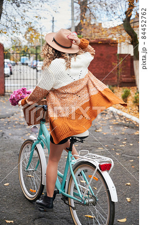 Rear view of biker riding on sidewalk by bright women's bicycle, holding hat on head with her right hand. On the background brick wall with gate, blurred silhouettes of cars. 84452762