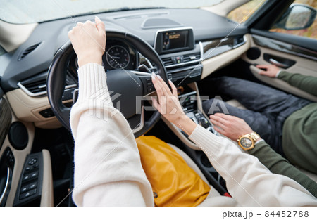 Close-up of fragile female hands driving elite stylish car on the background of dashboard and windshield. Cropped view of men's hands and lower torso at the right. 84452788