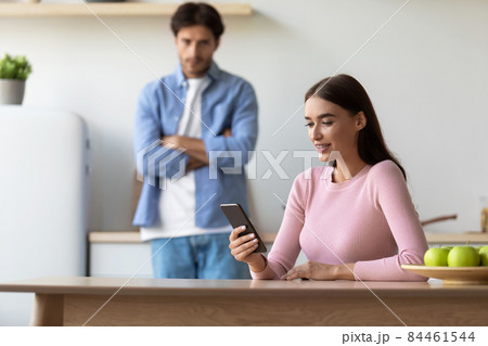 Smiling young european lady surfing in smartphone and ignores offended sad husband at kitchen 84461544