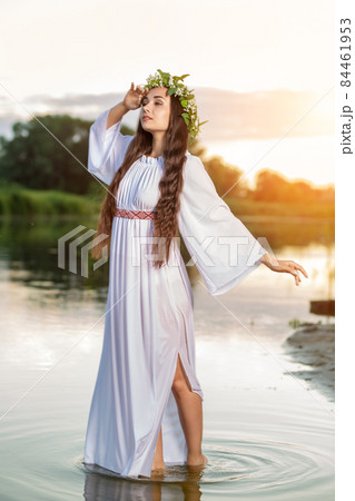 Beautiful black haired girl in white vintage dress and wreath of flowers standing in water of lake. Sun flare. Beautiful black haired girl in white vintage dress and wreath of flowers standing in water of lake. Sun flare. 84461953