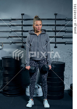 Beautiful young woman fitness instructor wearing a sports suit doing an exercise lunges with dumbbells on a gray background Beautiful young woman fitness instructor wearing a sports suit doing an exercise lunges with dumbbells on a gray background 84485391