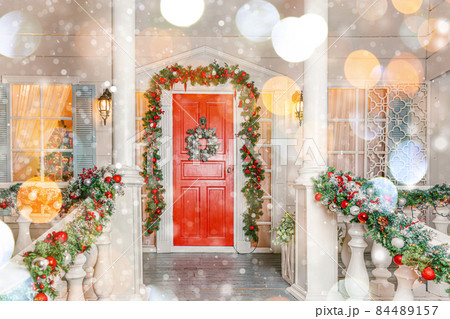 Christmas porch decoration idea. House entrance with red door decorated for holidays. Red and green wreath garland of fir tree branches and lights on railing. Christmas eve at home. 84489157