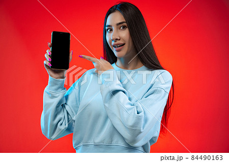 Cropped portrait of young joyful girl pointing at phone screen isolated over red background 84490163