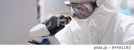 Scientist in protective suit and mask looks through microscope Scientist in protective suit and mask looks through microscope 84491183