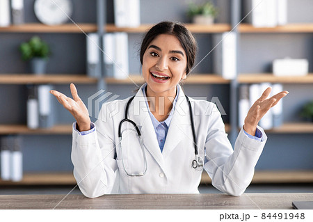 Smiling millennial attractive indian woman doctor in white coat gesturing and talking at workplace in clinic 84491948