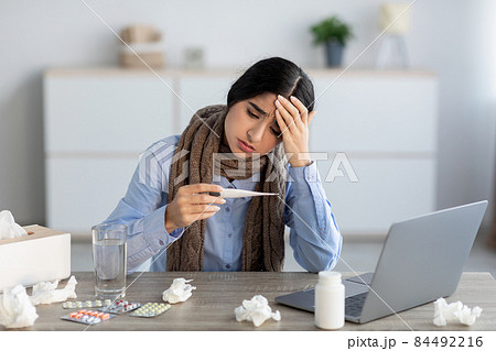 Sad young hindu woman manager suffering from headache, colds and flu looks at thermometer 84492216