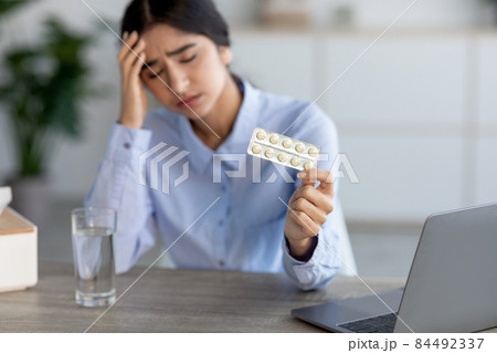 Despaired sad young indian lady suffering from pain, migraine and illness in workplace with laptop holds pills 84492337