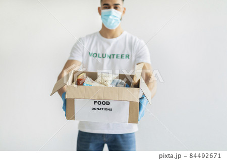 Food donations. Millennial arab man in volunteer t-shirt and medical mask, showing box with canned food and packages 84492671