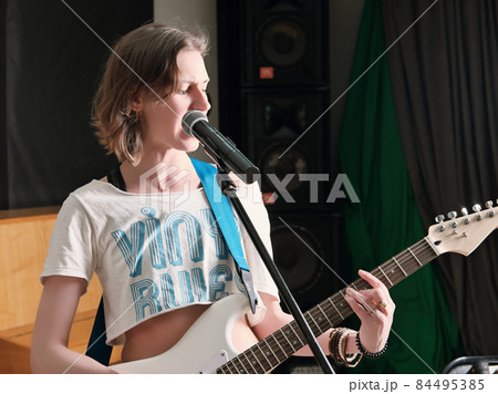 young woman musician playing guitar in studio during rehearsal. rock musician smiling and standing next to a microphone 84495385