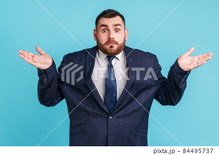 Puzzled clueless man with beard wearing official style suit spreading hands and shrugging shoulders, not sure and indifferent in some questions. Indoor studio shot isolated on blue background. 84495737