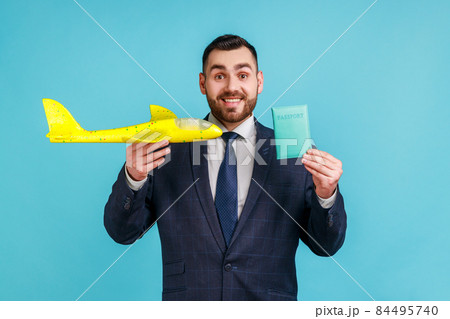 Positive man wearing official style suit holding passport and paper airplane, looking at camera with happy expression, satisfied by journey. Indoor studio shot isolated on blue background. Positive man wearing official style suit holding passport and paper airplane, looking at camera with happy expression, satisfied by journey. Indoor studio shot isolated on blue background. 84495740