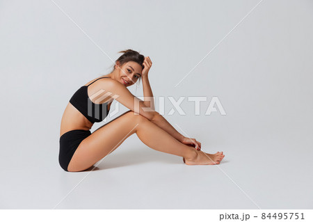 Portrait of young beautiful slim girl in black underwear sitting on floor isolated over gray studio background. Natural beauty concept. Portrait of young beautiful slim girl in black underwear sitting on floor isolated over gray studio background. Natural beauty concept. 84495751
