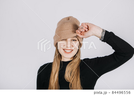 Unaltered candid emotional portrait of young happy blonde long hair woman covering eyes with brown knitted beanie hat. Portrait of playful smiling woman in knitted winter cap Unaltered candid emotional portrait of young happy blonde long hair woman covering eyes with brown knitted beanie hat. Portrait of playful smiling woman in knitted winter cap 84496556