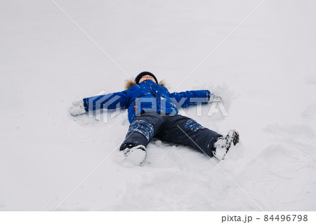 Best Outdoor Winter Activities for kids. Little kid boy in blue winter jacket making snow angel, laying down on snow in winter backyard. Active outdoors leisure with children in winter. 84496798