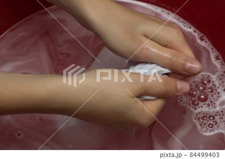 The girl erases a children's white T-shirt in a red basin from stains, cleans from stain remover, close-up, hands, white background 84499403