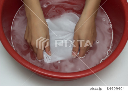 The girl erases a children's white T-shirt in a red basin from stains, cleans from stain remover, close-up, hands, white background The girl erases a children's white T-shirt in a red basin from stains, cleans from stain remover, close-up, hands, white background 84499404