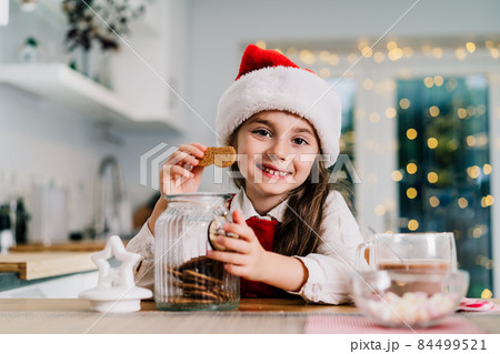 Happy smiling cute little girl kid in Santa Claus hat sitting on the decorated for winter holidays modern kitchen and eating gingerbread cookies from the jar with cocoa drink. Festive season. Happy smiling cute little girl kid in Santa Claus hat sitting on the decorated for winter holidays modern kitchen and eating gingerbread cookies from the jar with cocoa drink. Festive season. 84499521