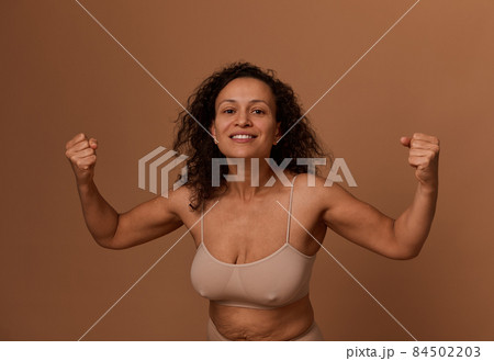 Delighted smiling dark-haired curly woman in beige underwear, clenching her fists, smiling with a toothy smile, looking at the camera. Body positivity, concept of combating oppression of women rights Delighted smiling dark-haired curly woman in beige underwear, clenching her fists, smiling with a toothy smile, looking at the camera. Body positivity, concept of combating oppression of women rights 84502203