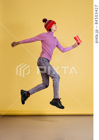 Full-length portrait of jouful young woman in winter warm red hat jumping isolated over yellow background 84503427