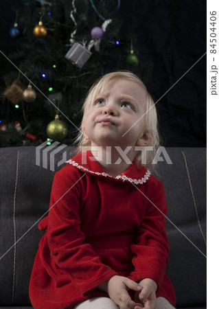 Little blonde girl in a red dress sits on the sofa and dreams against the background of a Christmas tree and a black wall. Christmas Eve. New Year Little blonde girl in a red dress sits on the sofa and dreams against the background of a Christmas tree and a black wall. Christmas Eve. New Year 84504406