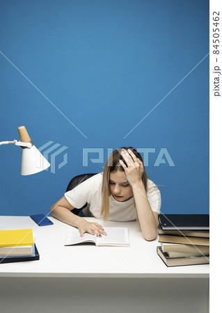 Tired young student woman in white casual clothes sitting at the table reading the book in library of university or college. Sitting and reading on blue background. Studying. 84505462