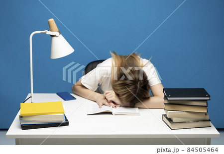 Tired student girl with glasses sleeping on the books in the library. Student studying hard exam and sleeping on books on a blue background. 84505464