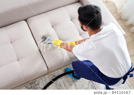 Close-up of housekeeper holding modern washing vacuum cleaner and cleaning dirty sofa with professionally detergent. Professional springclean at home concept 84510167