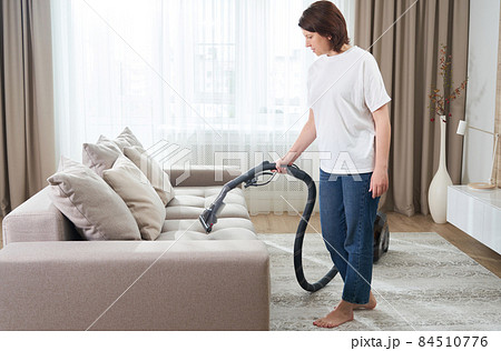 young woman in white shirt and jeans cleaning carpet under sofa with vacuum cleaner in living room, copy space. Housework, cleanig and chores concept young woman in white shirt and jeans cleaning carpet under sofa with vacuum cleaner in living room, copy space. Housework, cleanig and chores concept 84510776