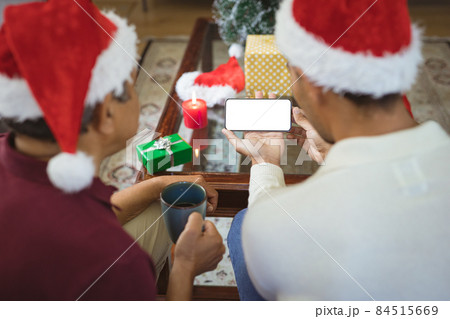 Biracial adult son and senior father in santa hats making smartphone christmas video call Biracial adult son and senior father in santa hats making smartphone christmas video call 84515669
