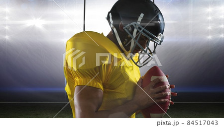 Side view of male american football athlete wearing helmet and yellow jersey holding ball at stadium 84517043